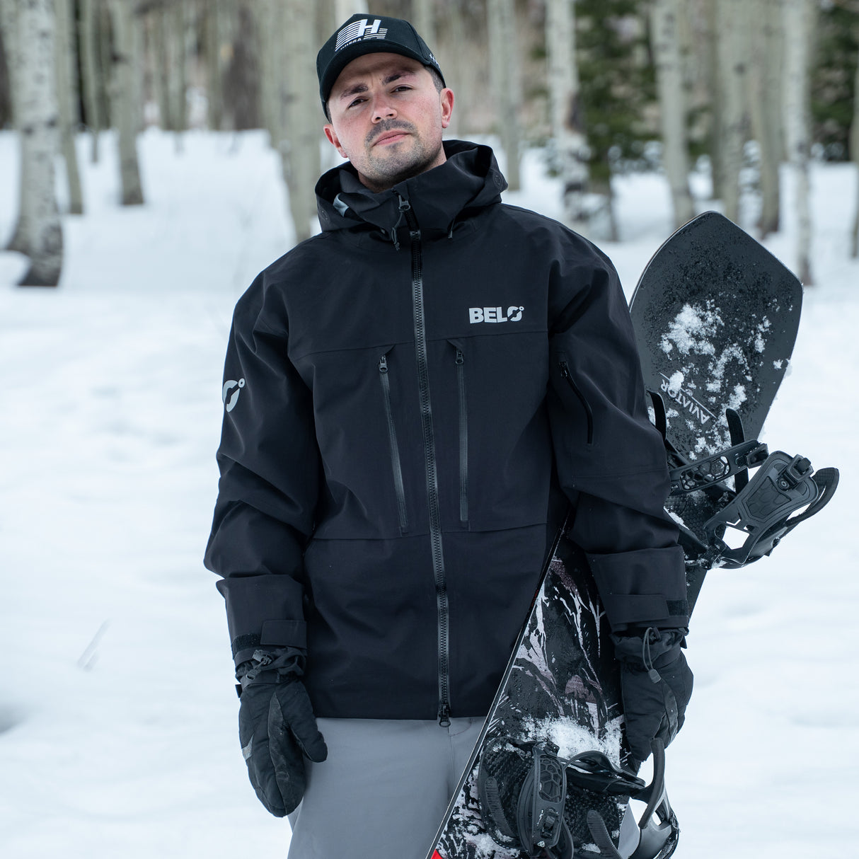 Man in black jacket holding a snowboard in a snowy forest